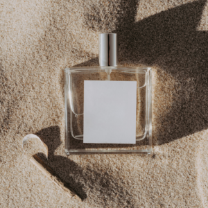 A clear glass bottle of men's fragrance with a blank label lying on sand, next to a piece of driftwood.